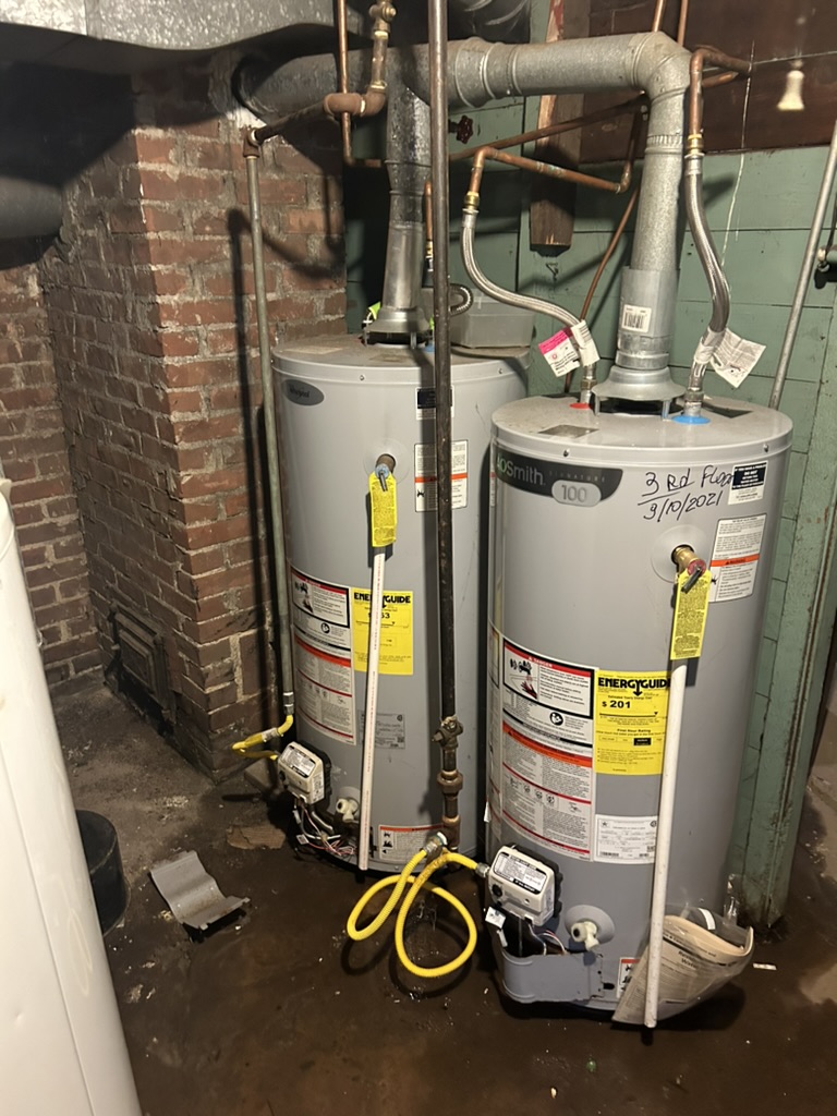D17F3AA7-9F8A-434B-9982-437E16ED2E5D_1_105_c Two residential gas water heaters installed side-by-side in a basement, connected to yellow flexible gas lines and galvanized venting. Labeled "3rd Floor 3/10/2021", the units show EnergyGuide labels and digital gas meters. Visible brick wall and utility piping surround the setup.