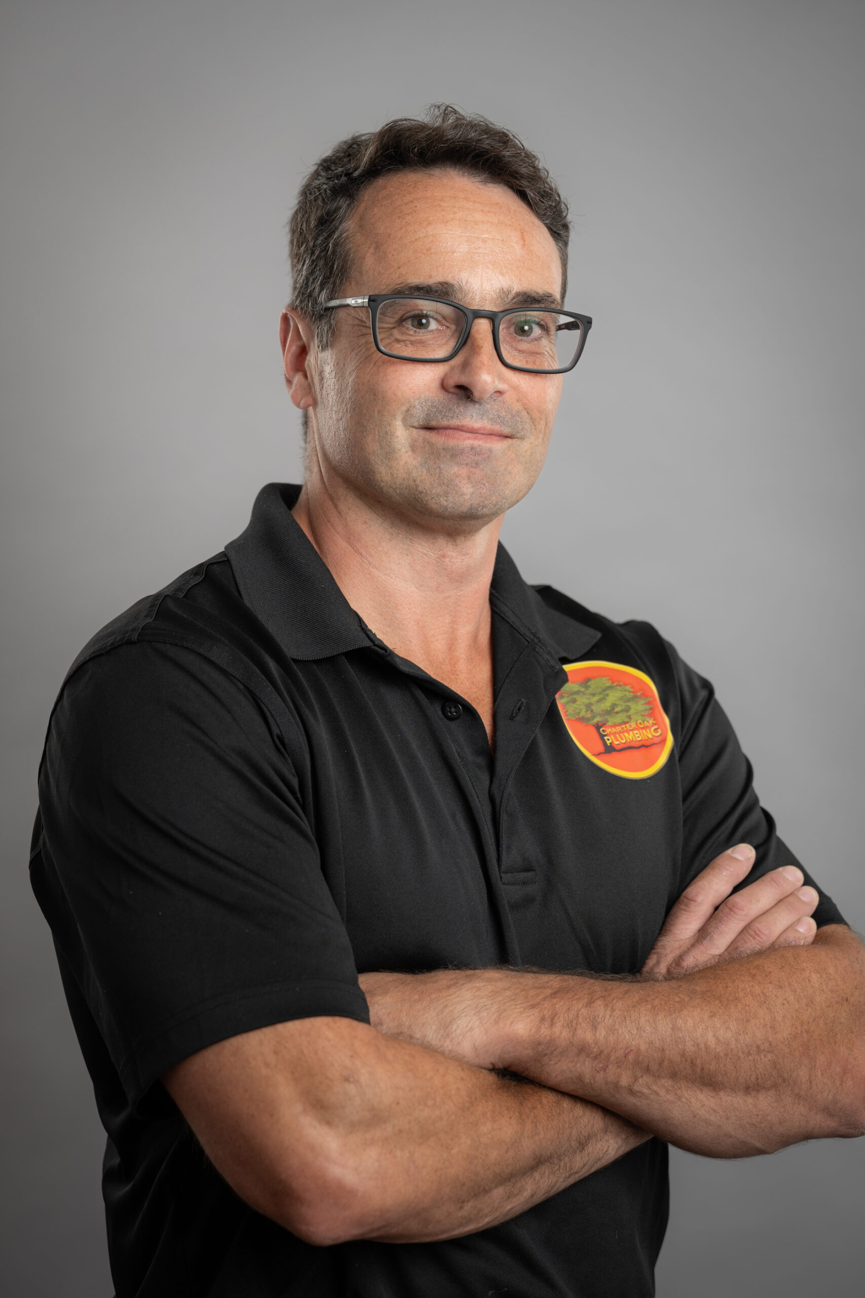 Headshote Paul, a confident and professional plumber, stands with arms crossed wearing a black polo shirt with the "Cherry Oak Plumbing" logo, set against a neutral gray background.