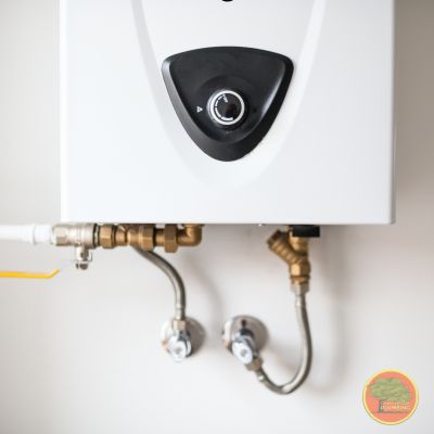 Tanklerss WH A close-up view of a modern tankless water heater mounted on a wall. The unit features a black central dial for temperature control. Below, connected water lines and valves—both metal and PVC—are neatly installed. The setup appears clean and professionally done. A small circular logo is visible in the bottom right corner.